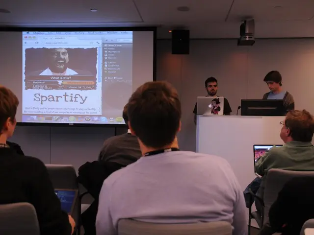 In this picture we can see a group of people where some are sitting on chairs and two men are...