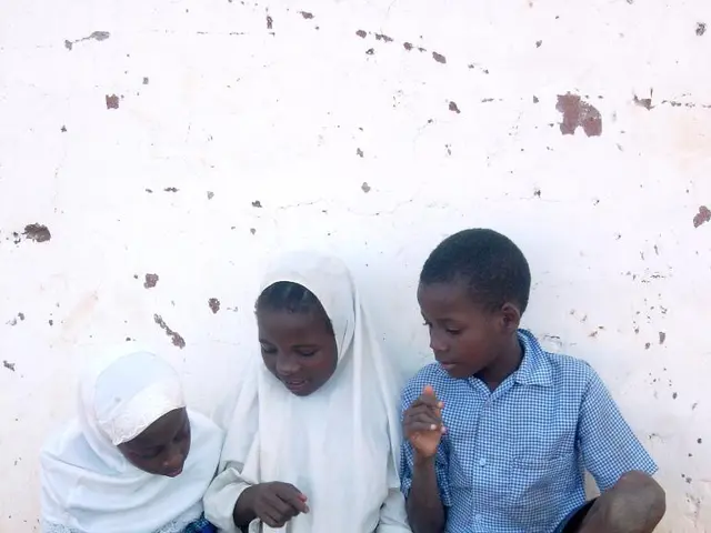 In the center of the picture there are three kids sitting on the ground. In the foreground there is...